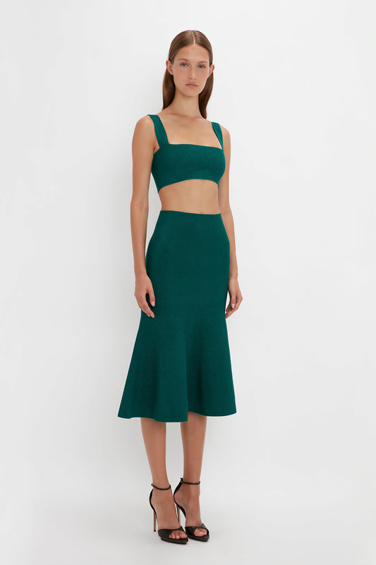 A woman stands against a plain white background, wearing a green crop top and a matching VB Body Flared Skirt In Lurex Green with a flared silhouette. She has long, straight hair and is posing with a neutral expression. The ensemble is from Victoria Beckham.