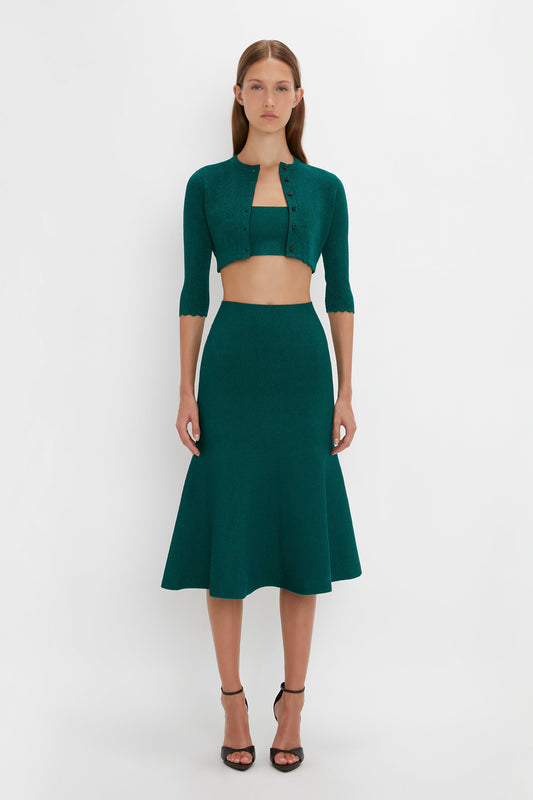A woman is standing against a plain backdrop, wearing the Victoria Beckham VB Body Cropped Cardi In Lurex Green, a green three-piece outfit consisting of a cropped cardigan with pointelle stitch detailing, a crop top, and a flared skirt, along with black heels.