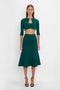 A woman is standing against a plain backdrop, wearing the Victoria Beckham VB Body Cropped Cardi In Lurex Green, a green three-piece outfit consisting of a cropped cardigan with pointelle stitch detailing, a crop top, and a flared skirt, along with black heels.