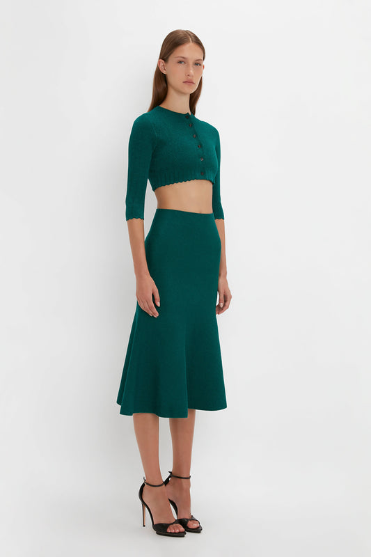A person stands against a plain background wearing a green, long-sleeved, VB Body Cropped Cardi In Lurex Green by Victoria Beckham paired with a matching high-waisted, flared skirt and black high-heeled sandals.