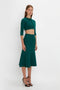 A person stands against a plain background wearing a green, long-sleeved, VB Body Cropped Cardi In Lurex Green by Victoria Beckham paired with a matching high-waisted, flared skirt and black high-heeled sandals.