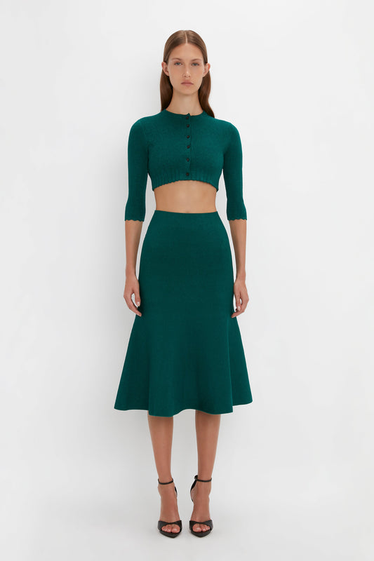 A woman stands against a plain white background, wearing a green VB Body Cropped Cardi In Lurex Green from Victoria Beckham, with pointelle stitch detailing and a matching flared green skirt, and black high-heeled sandals.
