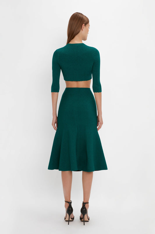 A woman with long hair is standing with her back to the camera, wearing a teal two-piece outfit consisting of a Victoria Beckham VB Body Cropped Cardi In Lurex Green and a flared skirt with pointelle stitch detailing, paired with black high-heeled sandals.