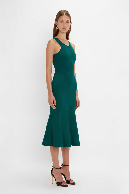 A woman stands facing slightly to the right, wearing a Victoria Beckham VB Body Sleeveless Dress In Lurex Green. The flared silhouette and black high-heeled sandals make this outfit a perfect addition to any new-season wardrobe. The background is plain white.