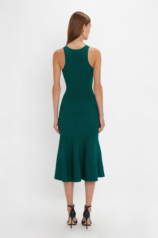 A woman with long brown hair is wearing a sleeveless, green VB Body Sleeveless Dress In Lurex Green by Victoria Beckham featuring a flared silhouette and black high-heeled shoes, shown from the back against a white background. Perfect for updating your new-season wardrobe.
