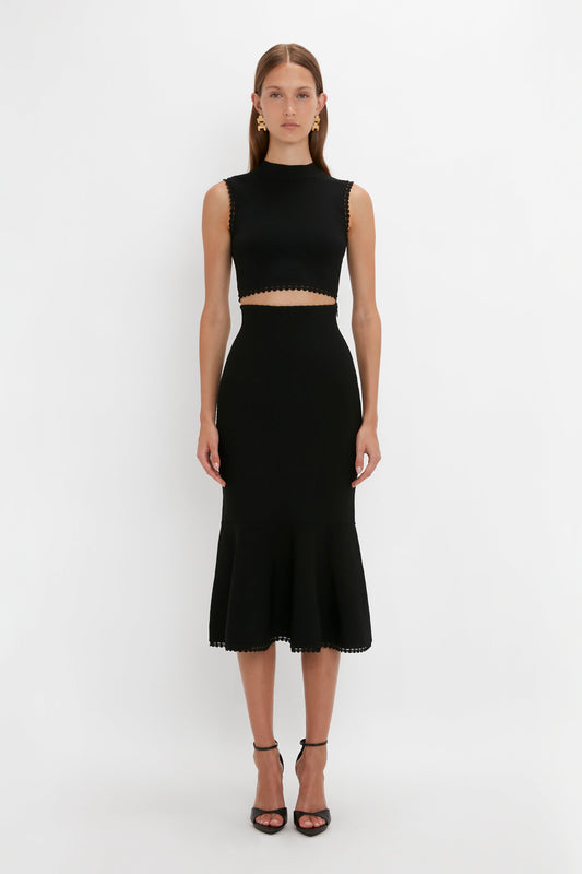 A woman stands against a plain background, showcasing the flattering flared silhouette of her sleeveless black crop top and matching Victoria Beckham VB Body Scallop Trim Flared Skirt In Black, paired perfectly with black high-heeled sandals.