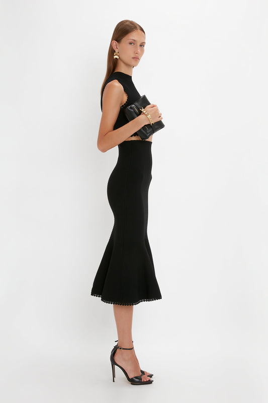 Woman in a black sleeveless dress and high-heeled sandals stands sideways, holding a black clutch. The Victoria Beckham VB Body Scallop Trim Flared Skirt In Black features a flattering flared silhouette, perfect for versatile styling. White background.