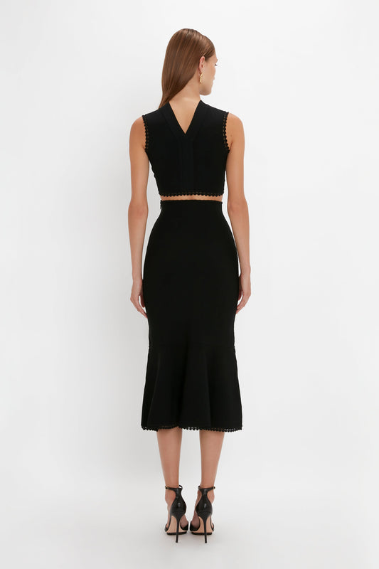 A woman is standing with her back to the camera, wearing a black sleeveless, v-neck dress with a flared silhouette and black high-heeled sandals, featuring the Victoria Beckham VB Body Scallop Trim Flared Skirt In Black.