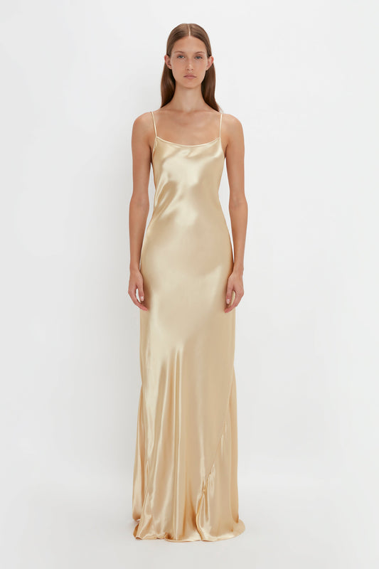 Person wearing a long, sleeveless, Victoria Beckham Exclusive Floor-Length Cami Dress In Gold with thin straps standing against a plain white background, evoking an elegant 1990s-inspired camisole-slip dress vibe.