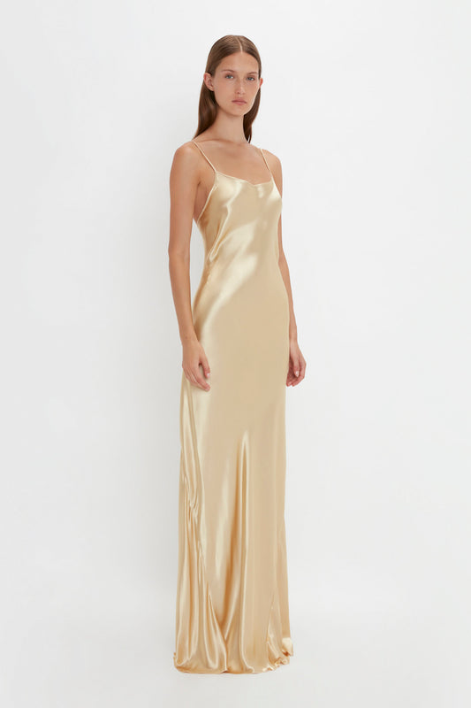 Person standing against a plain white background wearing the Victoria Beckham Exclusive Floor-Length Cami Dress In Gold with thin spaghetti straps, evoking a chic 1990s-inspired vibe.