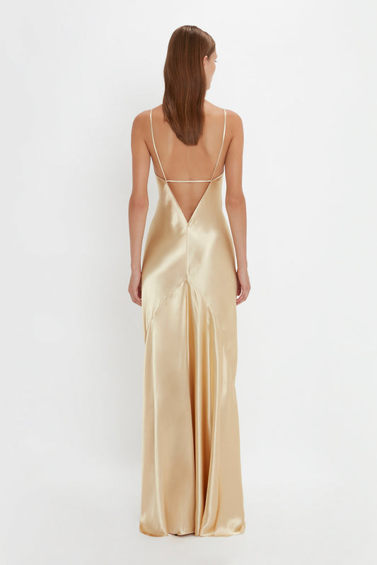 A woman wearing the Victoria Beckham Exclusive Floor-Length Cami Dress In Gold with thin straps, seen from behind against a plain white background, exudes a 1990s-inspired elegance.