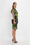 A woman in a green and black patterned Twist Shoulder Dress In Black Frost by Victoria Beckham, featuring draped pleat details, holds a black clutch and wears black high-heeled sandals, standing against a plain white background.