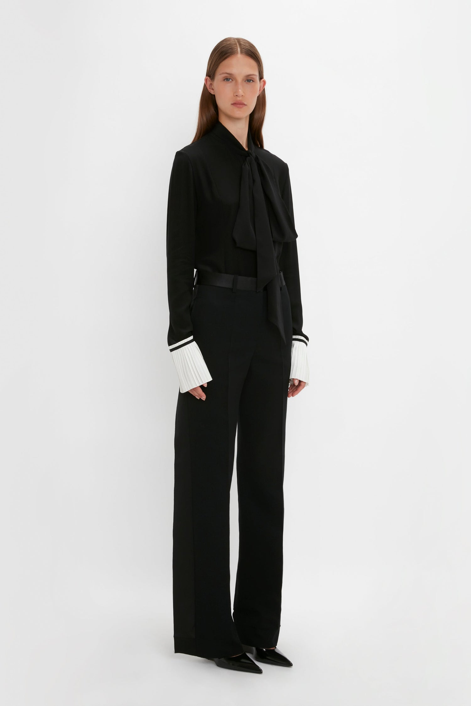 A person stands against a plain white background wearing a Pleat Cuff Blouse In Black by Victoria Beckham, long black pants, and pointed-toe black shoes.