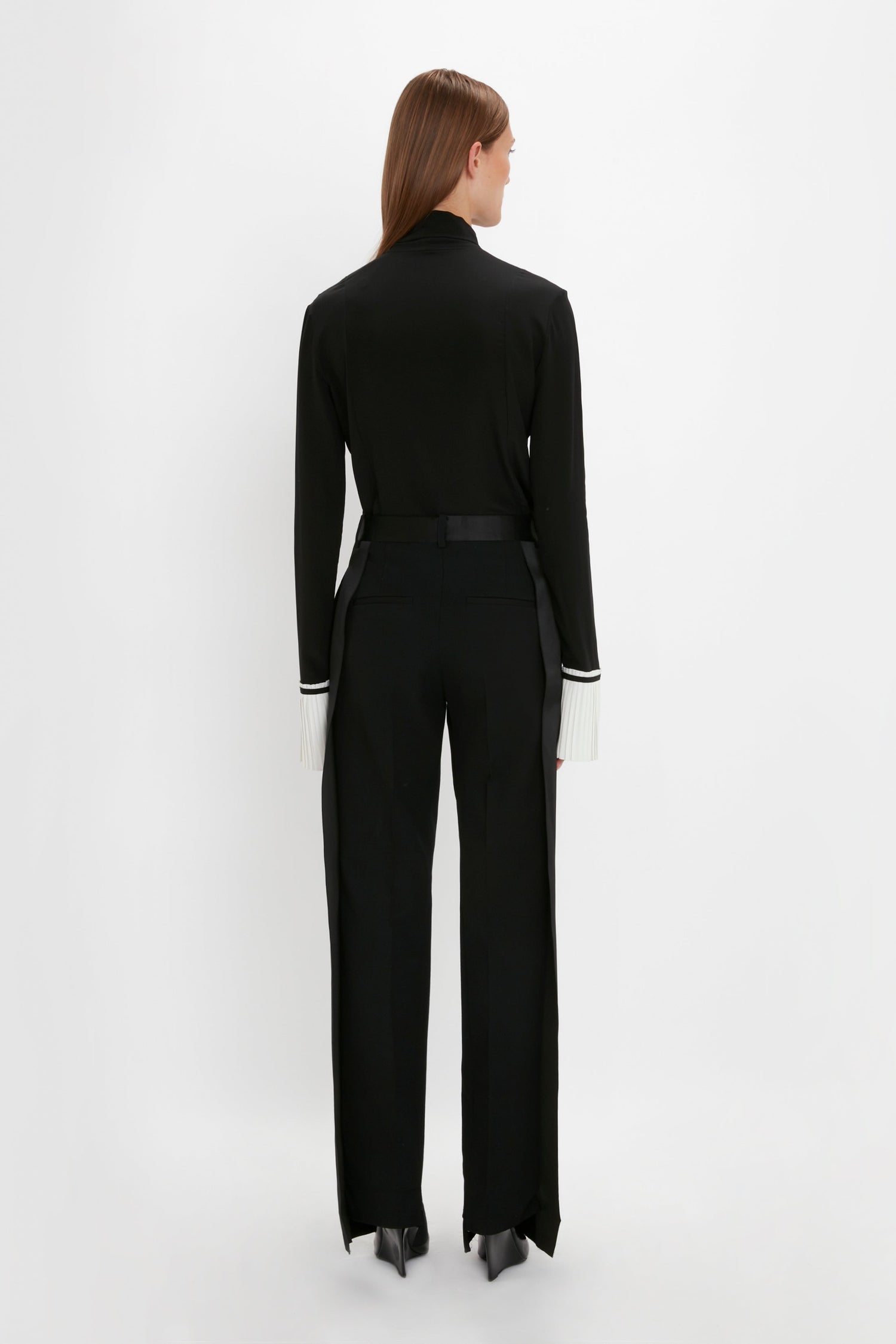 A person stands with their back to the camera, wearing a Pleat Cuff Blouse In Black and black trousers with white knife pleat cuffs by Victoria Beckham.