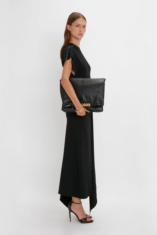 A woman in a crew-neck Short Sleeve Tie Detail Dress In Black by Victoria Beckham and Pointy Toe Stiletto Sandals poses sideways, holding a large black clutch with a golden clasp.