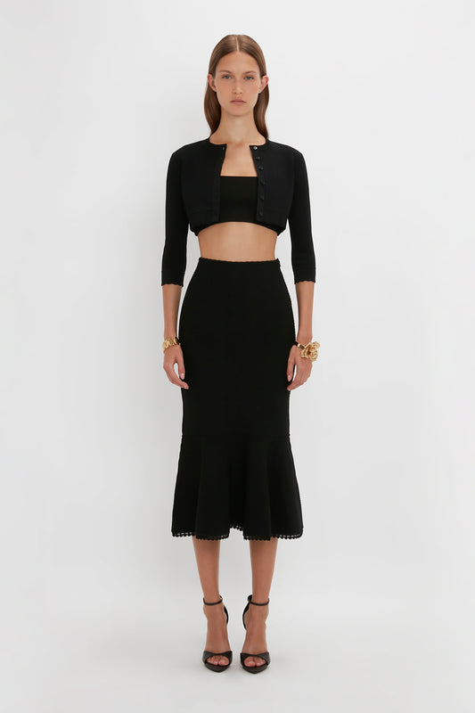 A person stands against a plain white background, wearing a black crop top and a black high-waisted skirt with a flared hem, exuding feminine sensibility. Complementing the look is a form-fitting VB Body Cropped Cardigan In Black by Victoria Beckham with three-quarter sleeves.