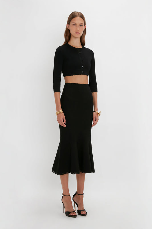 A woman stands against a plain white background, wearing a fitted black Victoria Beckham VB Body Cropped Cardigan In Black and a matching high-waisted skirt that flares at the hem, embodying a feminine sensibility. She is accessorized with gold bracelets and black high heels, highlighting her form-fitting silhouette.
