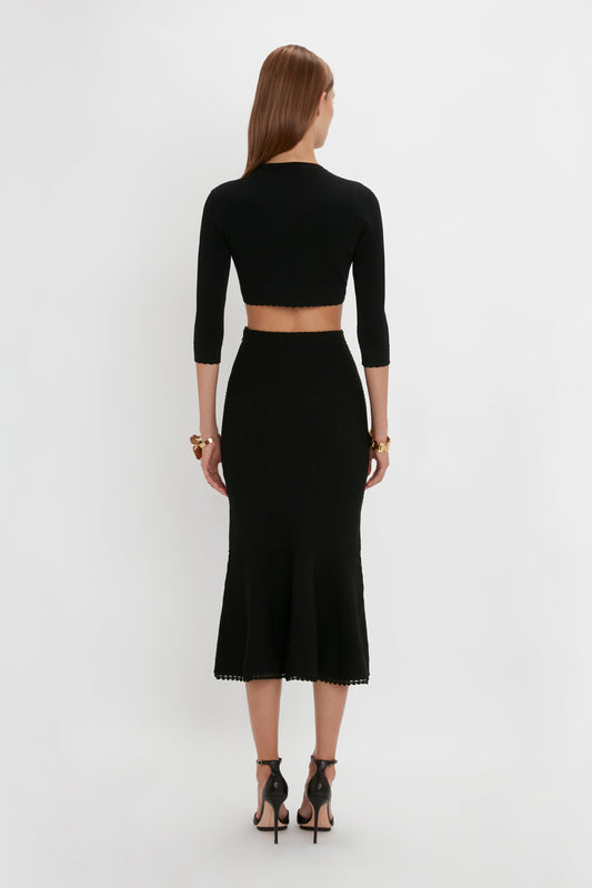 A person with long hair is standing with their back to the camera, wearing a black VB Body Cropped Cardigan In Black by Victoria Beckham and a matching midi skirt that perfectly highlights a form-fitting silhouette, complemented by black high-heeled sandals.