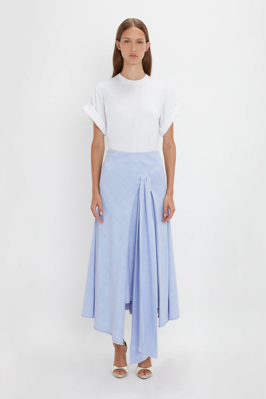 A woman stands against a white background wearing a white t-shirt and an Asymmetric Tie Detail Skirt In Frost by Victoria Beckham in light blue viscose crepe fabric with open-toe sandals, epitomizing modern femininity. She has straight, long brown hair.