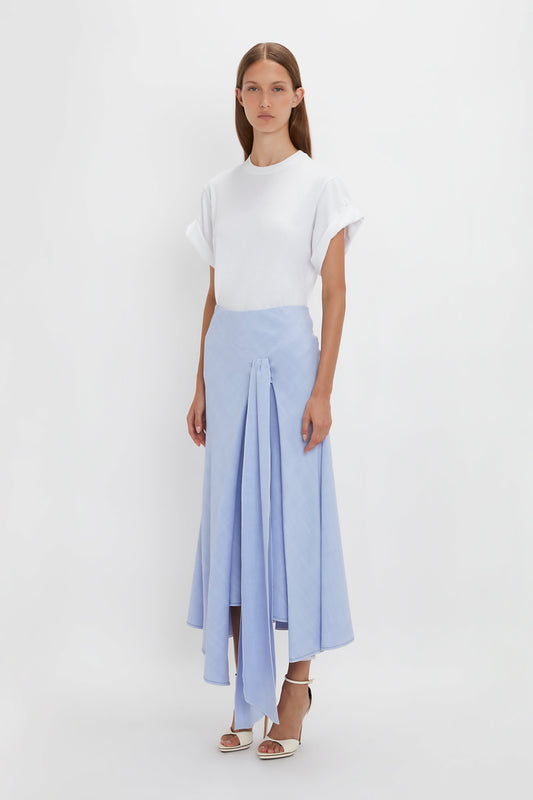 A woman stands against a plain white background wearing a white T-shirt and a light blue Asymmetric Tie Detail Skirt In Frost from Victoria Beckham made from viscose crepe fabric, paired with white high-heeled sandals.