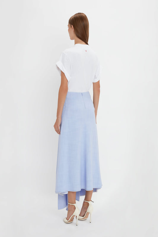 A woman stands with her back to the camera, embodying modern femininity in a white short-sleeve blouse and an Asymmetric Tie Detail Skirt In Frost by Victoria Beckham made from viscose crepe fabric. She completes the look with white high-heeled shoes, all set against a plain white background.