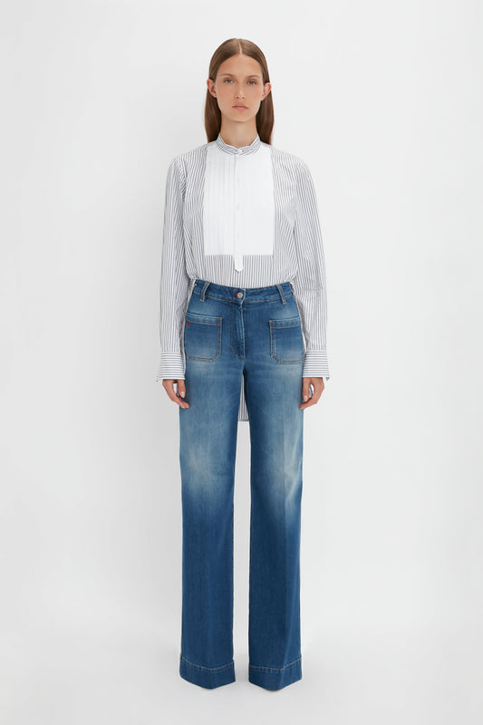 A person stands against a white background, wearing the Victoria Beckham Tuxedo Bib Shirt in Black and Off-White and faded blue wide-leg jeans, embodying relaxed menswear silhouettes.