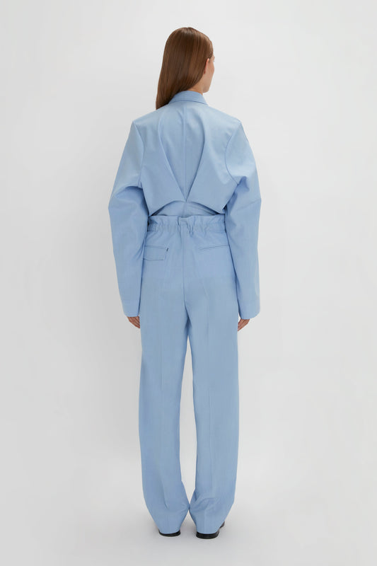 A person is standing with their back facing the camera, wearing an Oxford Blue oversized jacket and matching Gathered Waist Utility Trouser In Oxford Blue by Victoria Beckham with adjustable side tabs. The background is plain and white.