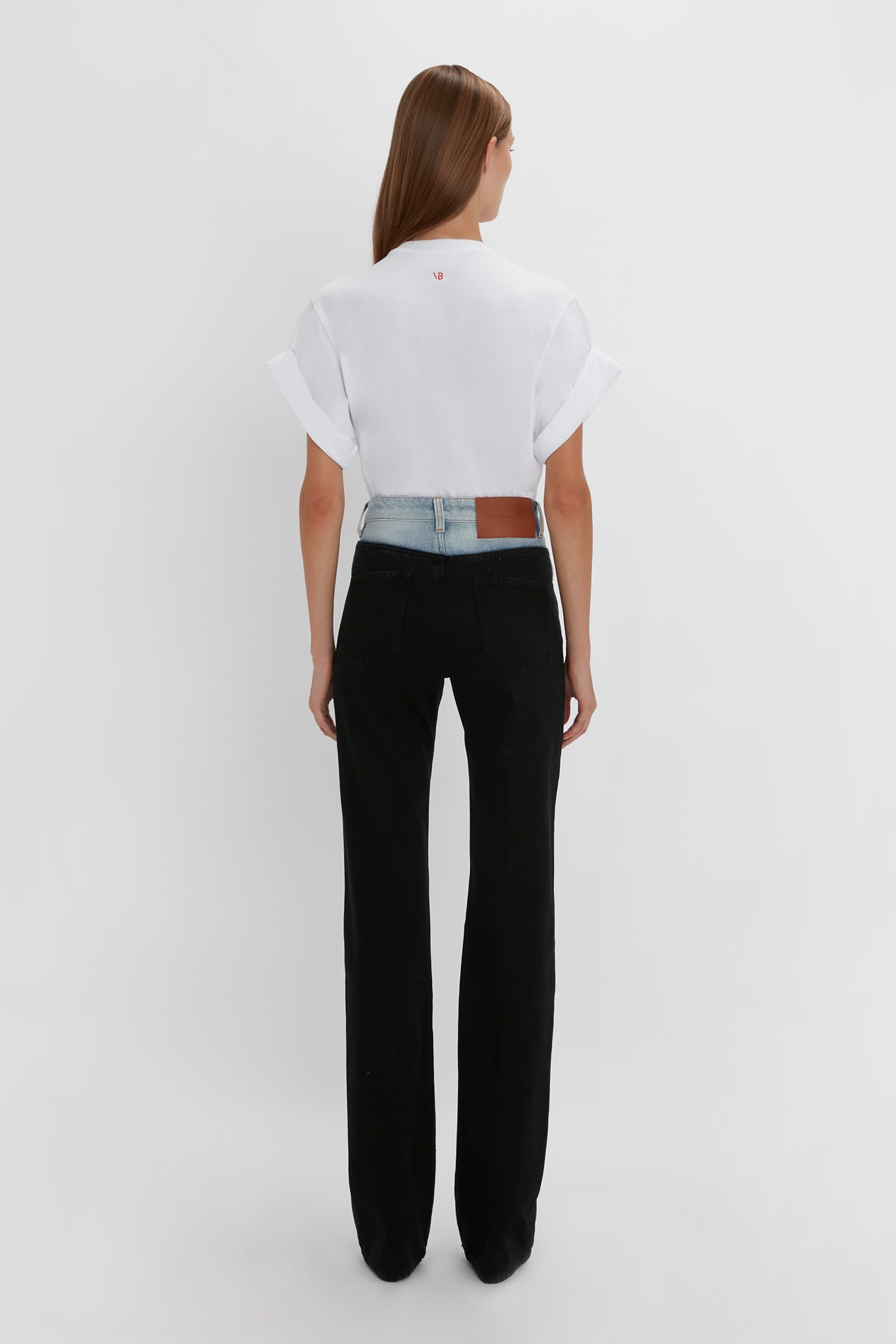 Woman standing with her back to the camera, wearing a white t-shirt and black Victoria Beckham Julia Jean In Contrast Wash on a plain background.