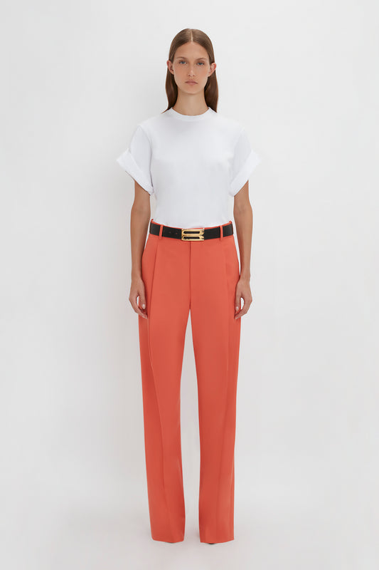 A person stands against a plain white background wearing a white T-shirt, Single Pleat Trouser In Papaya by Victoria Beckham with wide legs, and a black belt with a gold buckle.