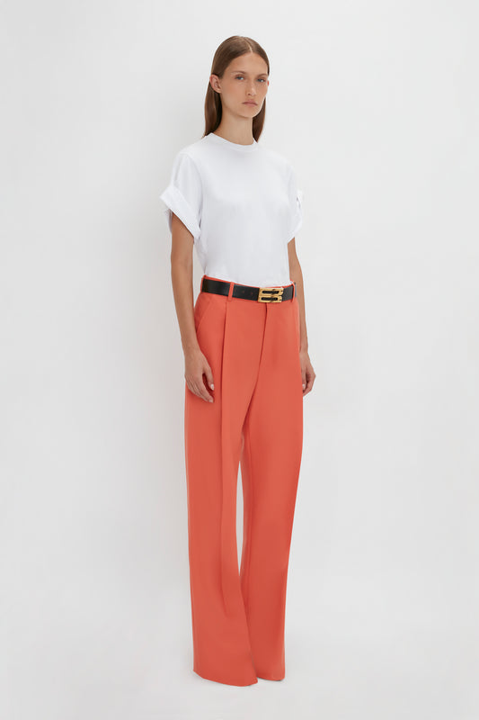 A person stands against a plain white background wearing a white t-shirt, Single Pleat Trouser In Papaya by Victoria Beckham with wide legs, and a black belt.