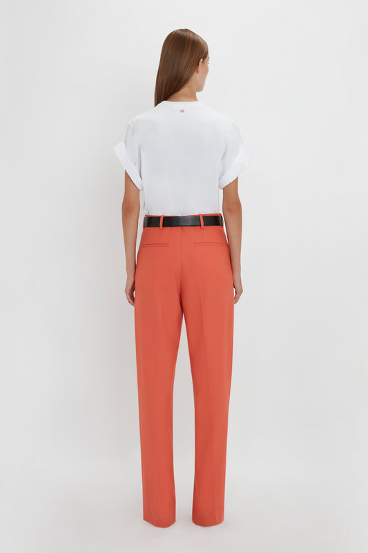 A person with long hair wearing a white short-sleeve top, black belt, and Single Pleat Trouser In Papaya by Victoria Beckham stands facing away from the camera.