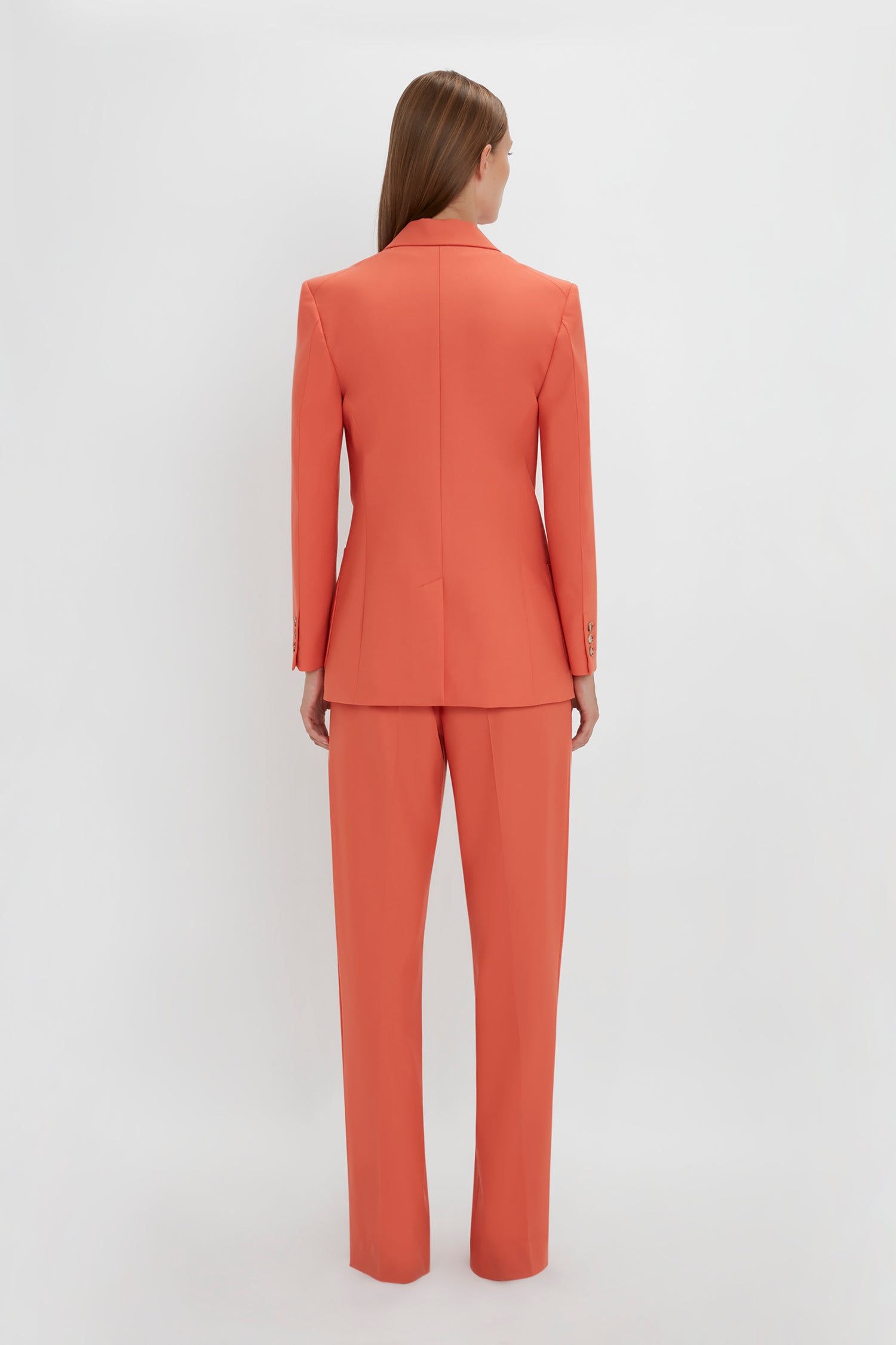 Rear view of a woman standing against a white background, wearing a Victoria Beckham tailored fit orange suit with straight-leg trousers.