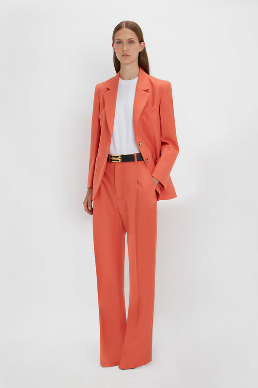 A person wearing a vibrant coral-colored pantsuit with Single Pleat Trouser In Papaya by Victoria Beckham, a white shirt, and a black belt stands against a plain white background.