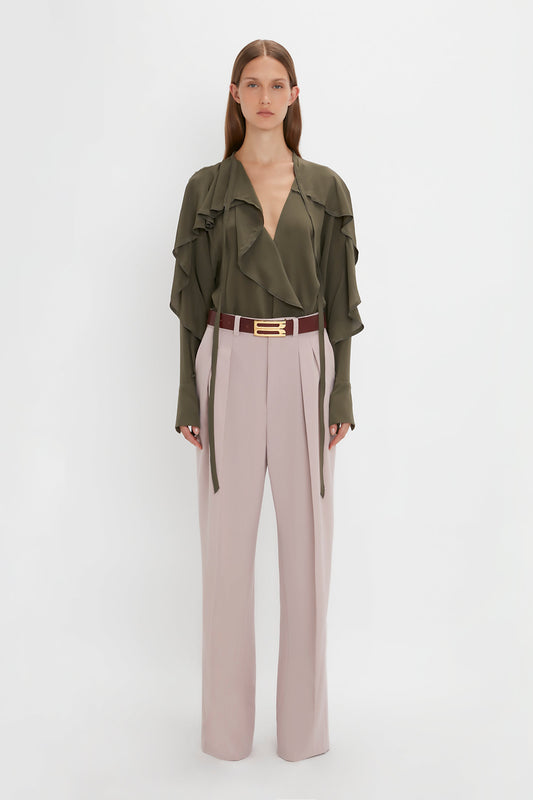Woman in an olive green ruffled blouse and Victoria Beckham Double Pleat Trouser In Rose Quartz, standing against a plain white background.