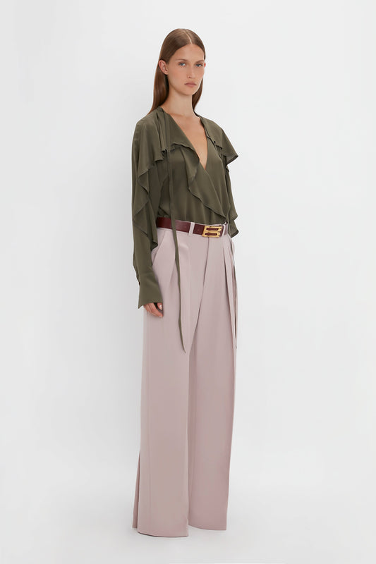 A woman stands against a plain background wearing a long-sleeve, v-neck **Tie Detail Ruffle Blouse in Oregano by Victoria Beckham** and pale pink double pleat trousers, accessorized with a burgundy belt.