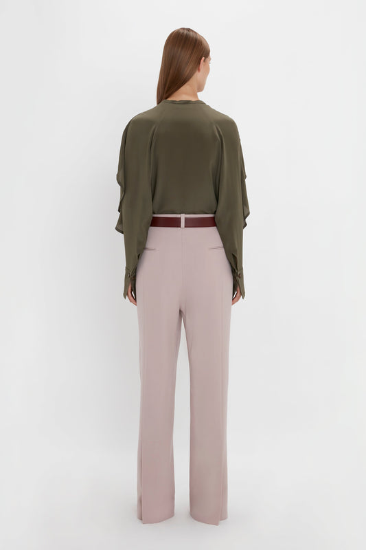 A person with long brown hair is standing with their back to the camera, wearing a Victoria Beckham Tie Detail Ruffle Blouse in Oregano and light pink double pleat trousers, secured with a burgundy belt.