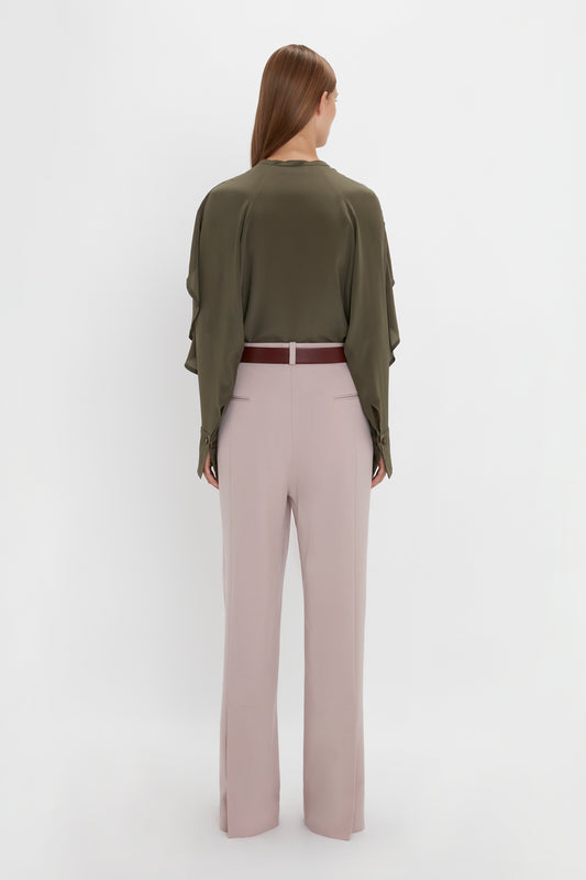A person stands with their back to the camera, wearing a dark green top and wide-leg, light pink Double Pleat Trouser In Rose Quartz by Victoria Beckham with a red belt, exuding a subtle masculine vibe.