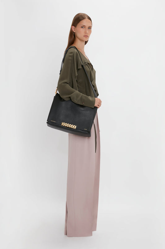 A person with long hair stands against a white background wearing a Tie Detail Ruffle Blouse in Oregano by Victoria Beckham and light pink Double Pleat Trousers, carrying a black handbag with gold accents on their shoulder.