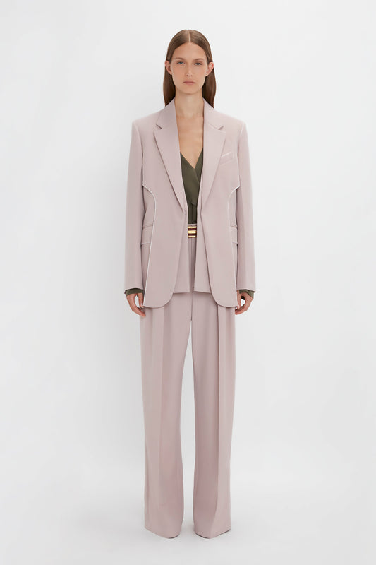 A person with long hair is wearing a light grey pantsuit featuring the Double Panel Front Jacket In Rose Quartz by Victoria Beckham, with a dark-colored top underneath, standing against a plain white background.
