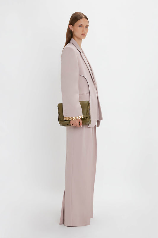 A woman with long hair, wearing a Victoria Beckham Double Panel Front Jacket In Rose Quartz and holding an olive green clutch, stands against a plain white background.
