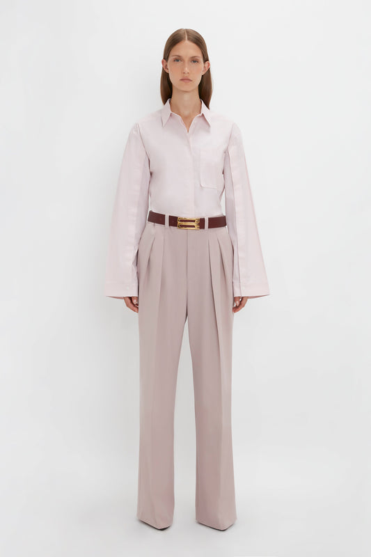 A person with long brown hair stands against a plain white background, wearing a Button Detail Cropped Shirt In Rose Quartz by Victoria Beckham, tan pleated wide-leg trousers, and a brown belt with a gold buckle.