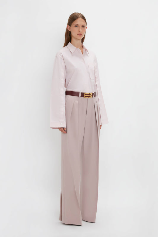A person stands against a white background wearing a Button Detail Cropped Shirt In Rose Quartz from Victoria Beckham, beige pleated wide-leg trousers, and a brown belt.
