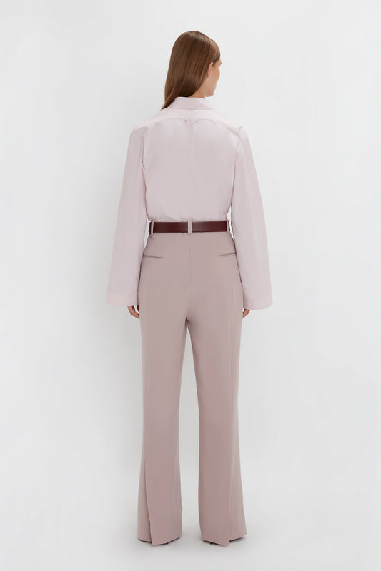 A person with long hair is standing facing away from the camera, wearing a Victoria Beckham Button Detail Cropped Shirt In Rose Quartz, tan trousers, and a brown belt.