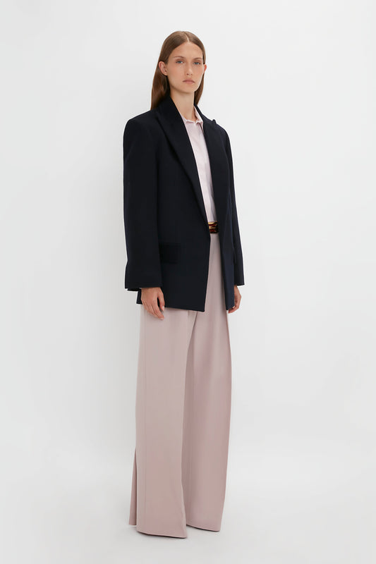 A person stands against a white background, exuding a contemporary feel in a Victoria Beckham Peak Lapel Jacket In Midnight over a white shirt and wide-legged light pink trousers.