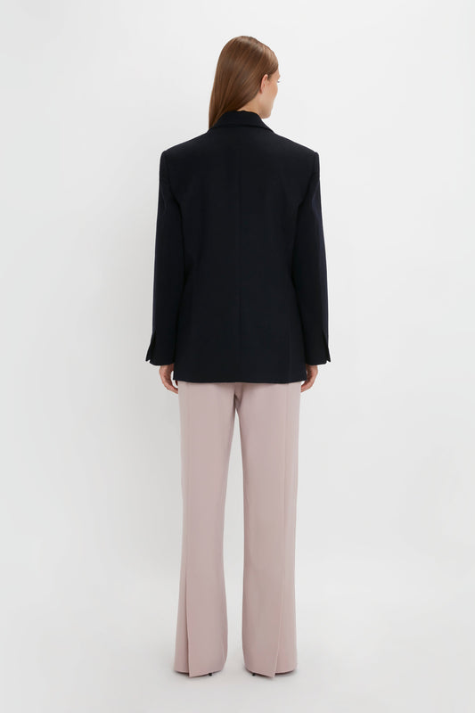 A person with long brown hair, wearing a black Peak Lapel Jacket In Midnight by Victoria Beckham and light pink pants, is standing facing away from the camera against a plain white background, giving the image a contemporary feel.
