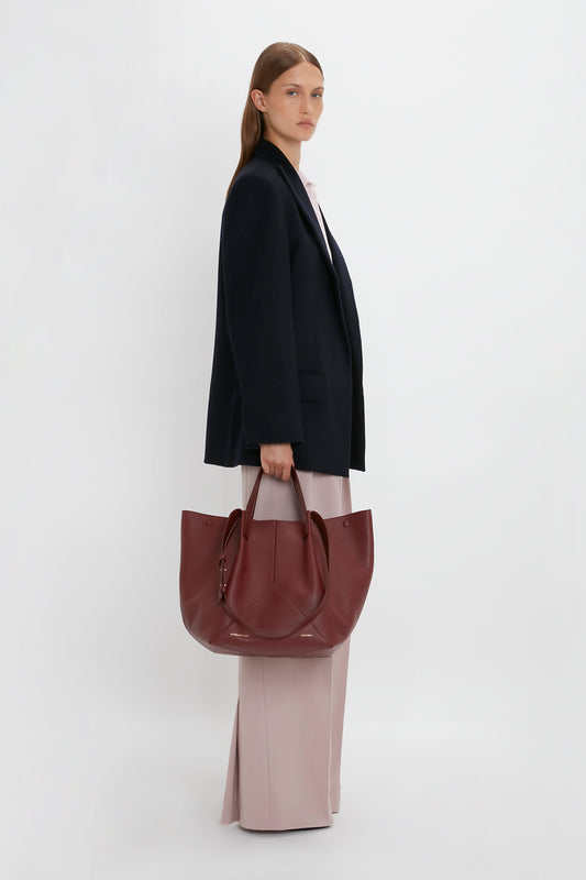 A woman with long hair, wearing a black Peak Lapel Jacket In Midnight by Victoria Beckham and light pink pants, is holding a large maroon handbag. She stands against a plain white background, exuding a contemporary feel reminiscent of Victoria Beckham's style.