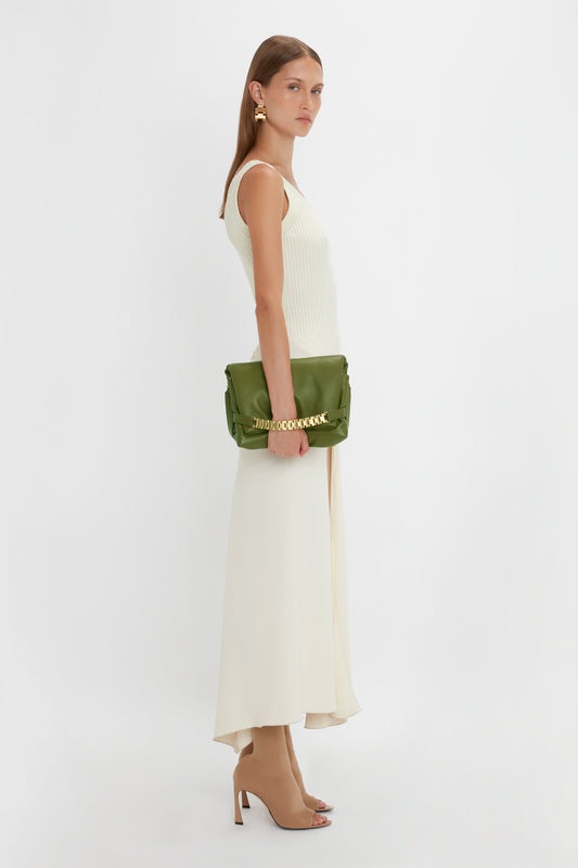 A woman stands against a plain white background wearing a sleeveless white dress and beige heels, holding a Puffy Chain Pouch With Strap In Khaki Leather from Victoria Beckham.