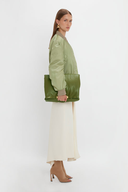 A woman in profile stands against a white backdrop, wearing a light green bomber jacket, a long white skirt, beige open-toe heels, and holding the Victoria Beckham Puffy Jumbo Chain Pouch In Khaki Leather.