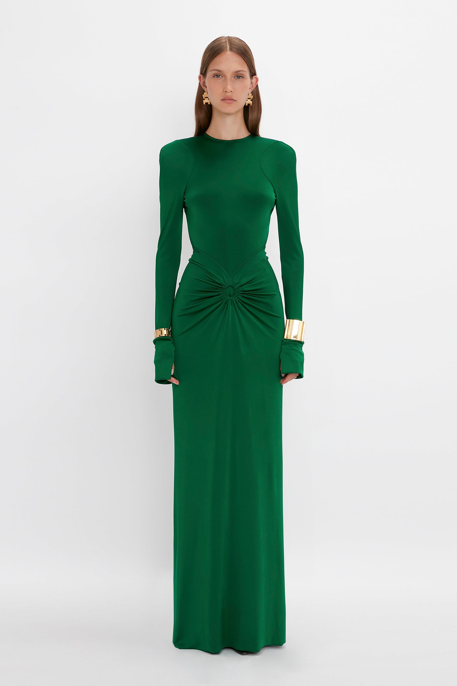 A woman in a long, emerald green dress stands against a plain white background. The Circle Detail Open Back Gown In Emerald by Victoria Beckham, made of body-sculpting jersey, features long sleeves, a high neckline, and a ruched detail at the waist. She wears gold cuff bracelets.