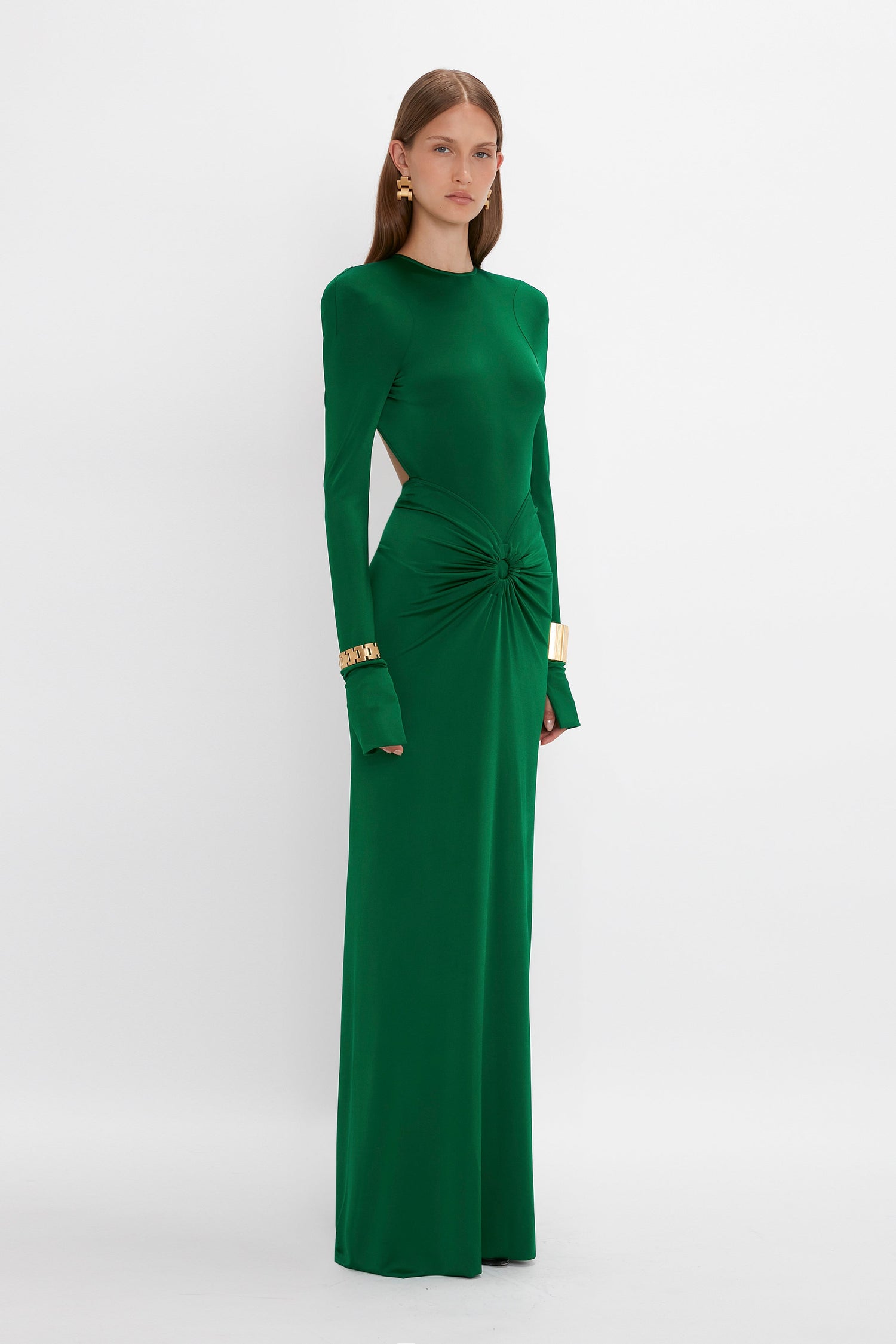A woman stands in a Victoria Beckham Circle Detail Open Back Gown In Emerald. The floor-length dress with side cutouts and gathered detailing at the waist is made from body-sculpting jersey fabric that accentuates her figure. She wears gold bracelets and long earrings. She has straight, long hair and a neutral expression.